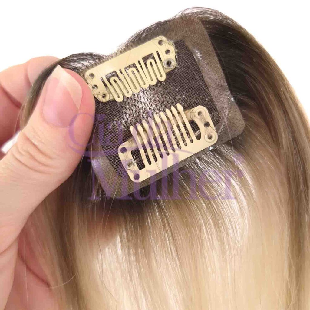 Aplique de franja de cabelo humano com base de tela marrom, projetado para disfarçar falhas. O cabelo apresenta um efeito ombre, começando em loiro escuro na raiz e transicionando para tons de loiro e caramelo. Duas presilhas de fixação beges estão visíveis na parte inferior da base. Uma mão, com os dedos polegar e indicador, segura a lateral superior esquerda do aplique.